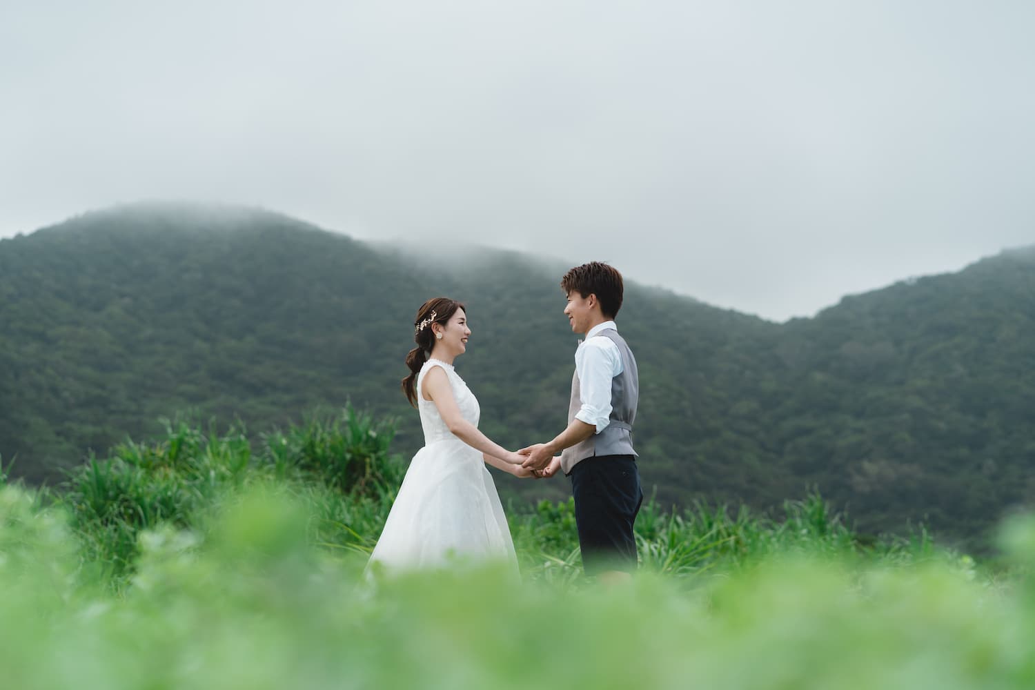 霧のかかった山を背景に手を取り合って立つ新郎新婦