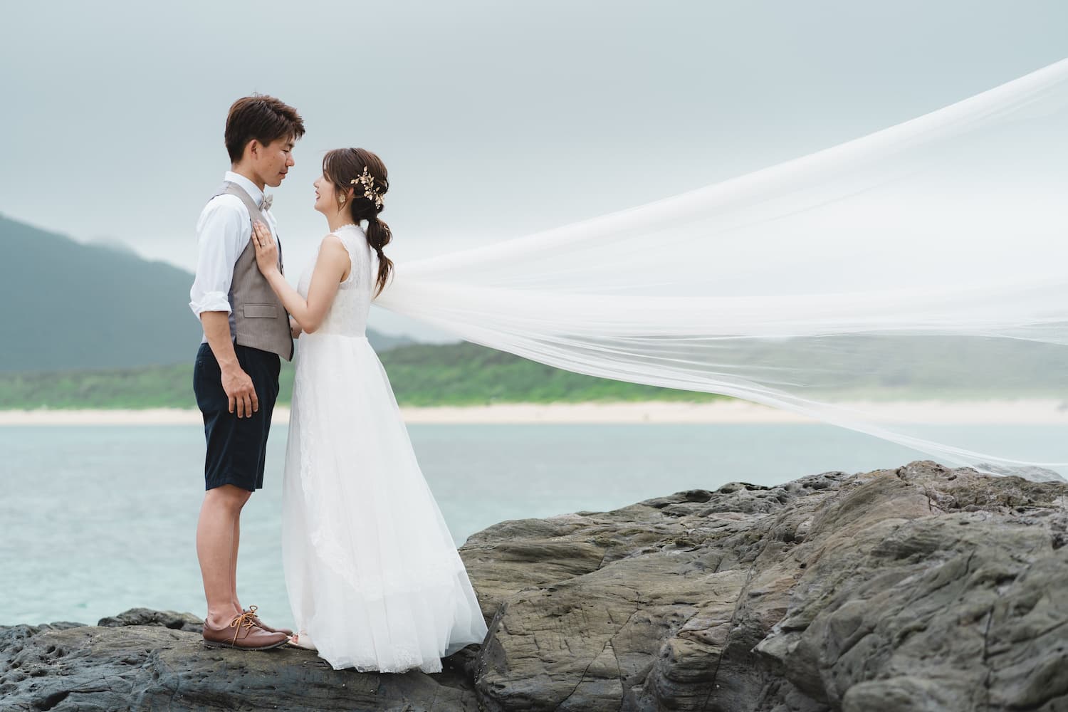 海にある岩場の上でベールを靡かせる新婦と新郎