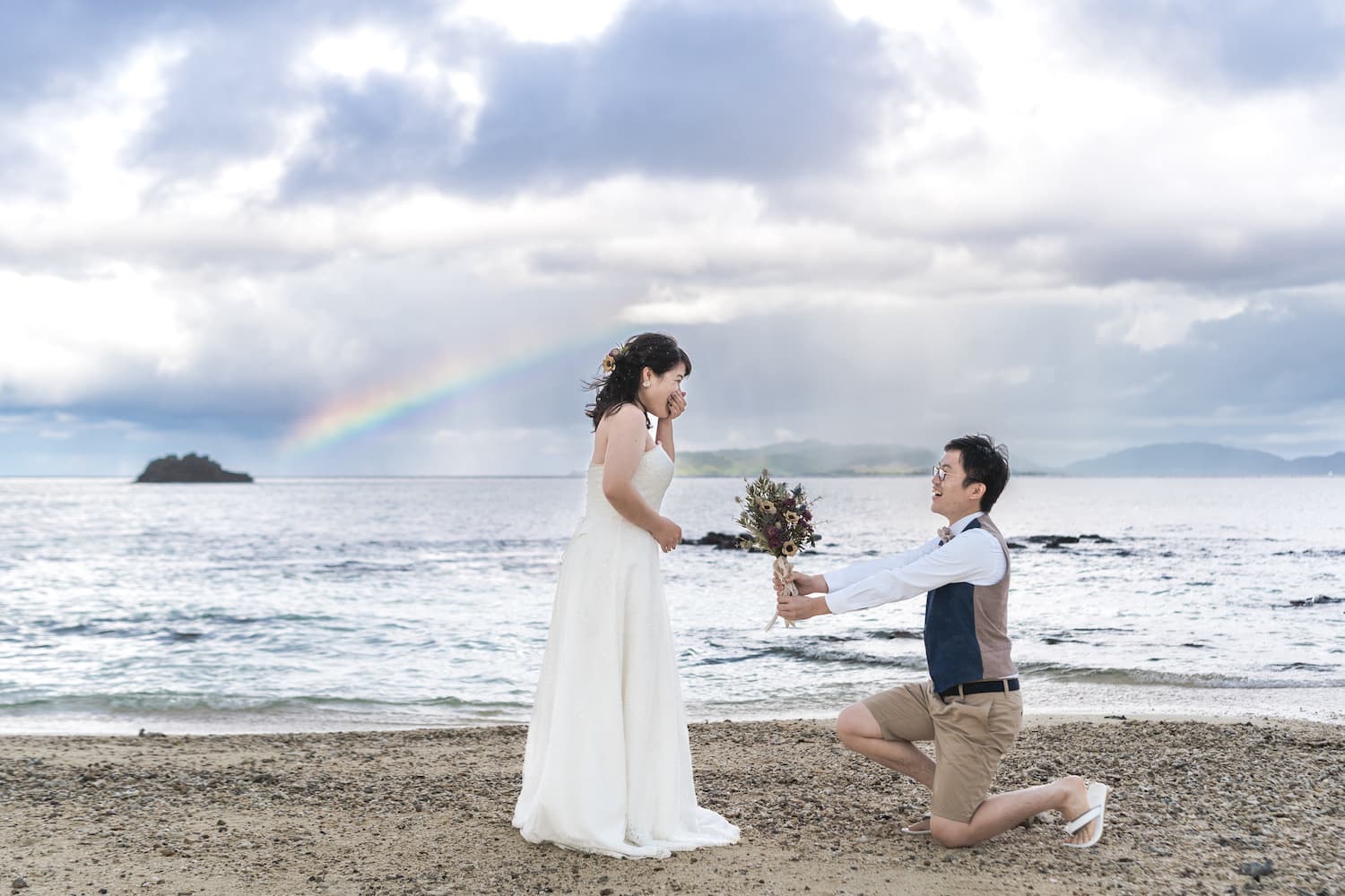 海に架かる虹を背景に新婦へブーケを差し出す新郎