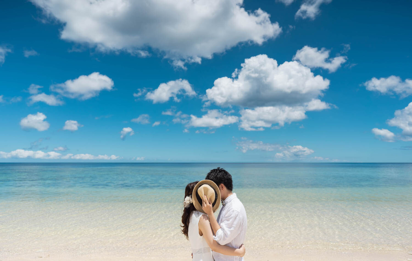 青空と海を背景に帽子で顔を隠す新郎新婦