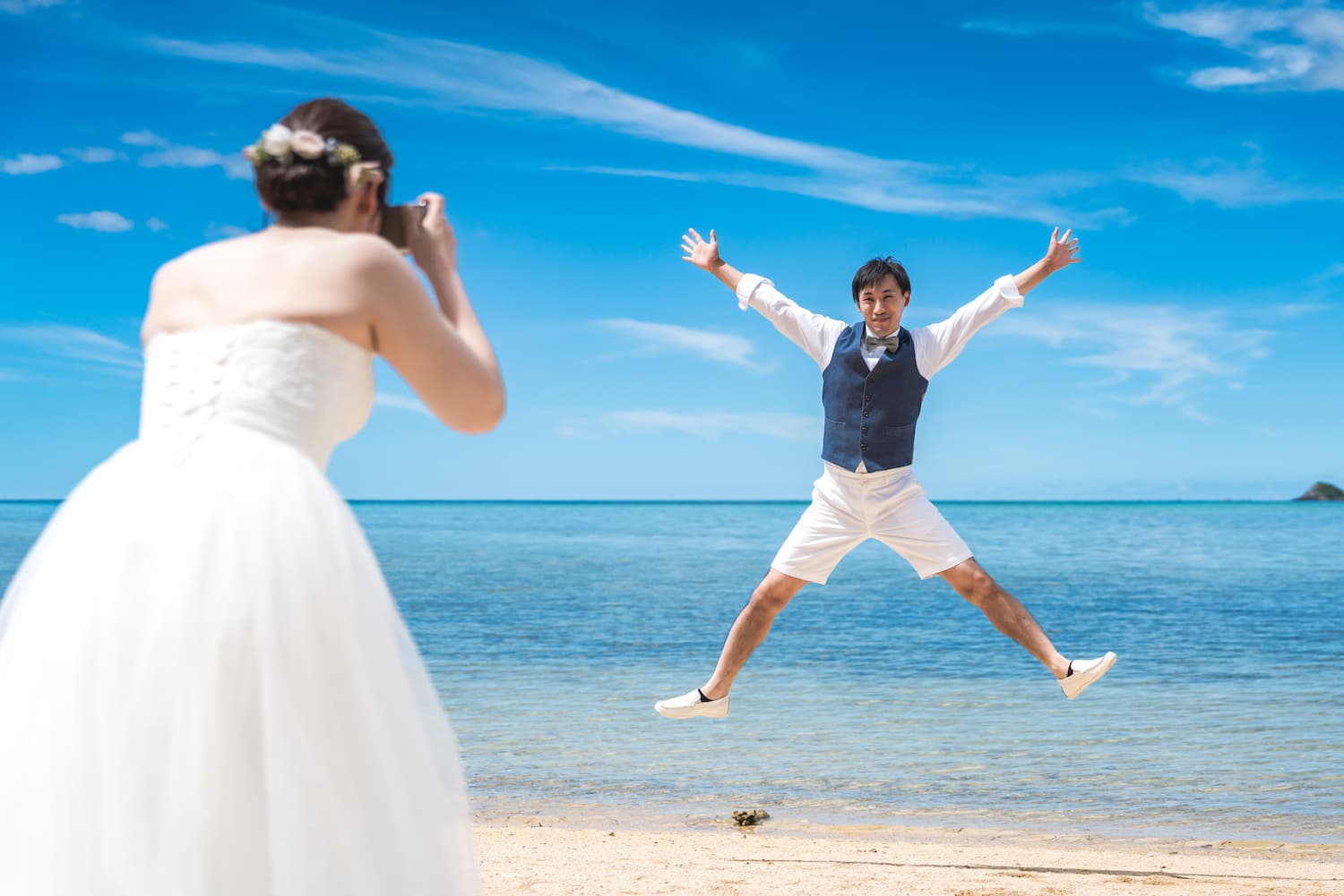 海を背景にジャンプする新郎とカメラを構える新婦