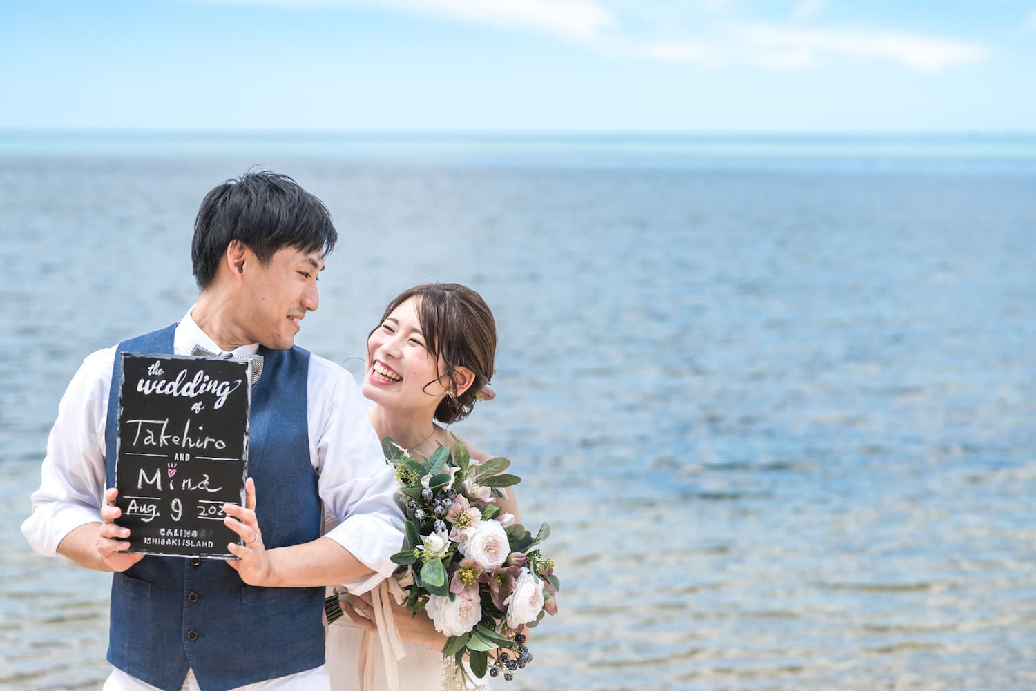 海を背景にボードを持って見つめ合う新郎新婦